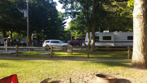 A campsite scene with parked RVs, a white car, and trees in a sunny outdoor setting.