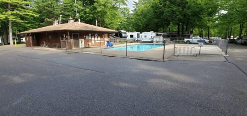A fenced pool area with a building nearby, surrounded by trees and parked RVs in a campground setting.