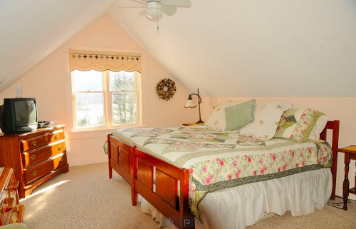 Cozy attic bedroom with a quilted bed, dresser, TV, and a window overlooking a scenic view.