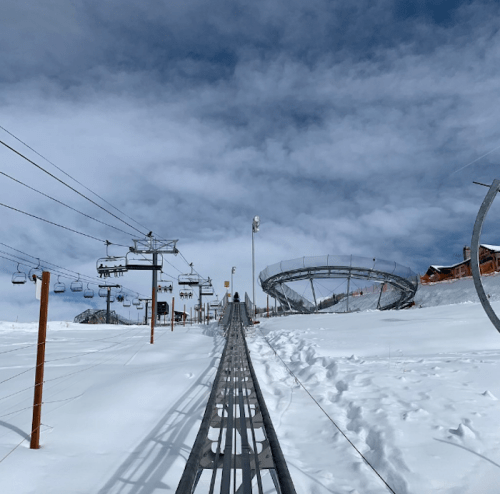 A snowy landscape featuring a ski lift, a winding track, and a cloudy sky in the background.