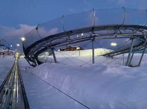 A snowy landscape featuring a roller coaster track and a lodge in the background, illuminated by evening lights.