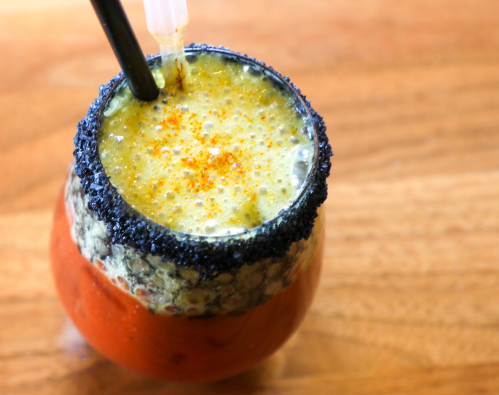 A colorful drink with a black salt rim, topped with spices, served in a glass on a wooden surface.