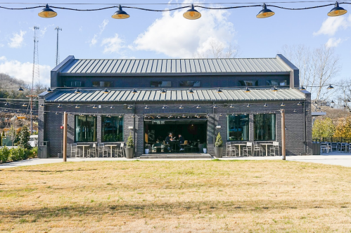 A modern building with a metal roof and large windows, surrounded by grass and string lights, under a blue sky.