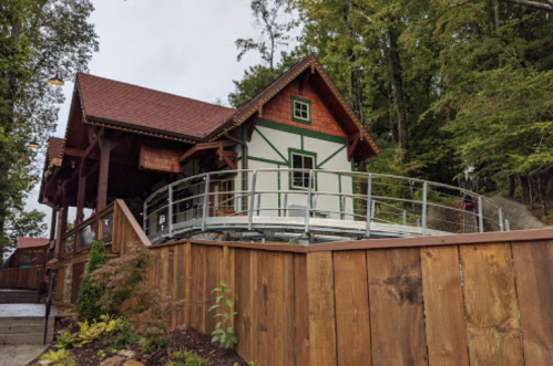 A charming wooden house with a red roof, surrounded by trees and a wooden fence, featuring a ramp for accessibility.