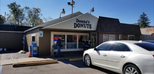A white car parked outside Donna's Donuts, with a person walking toward the entrance. The shop has an "OPEN" sign.