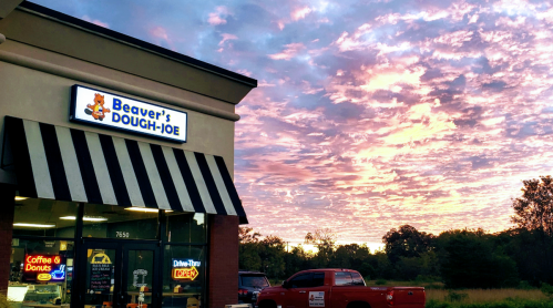 A donut shop named "Beaver's Dough-Joe" with a colorful sunset in the background and parked red trucks outside.