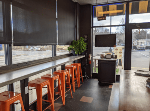 Interior of a café with a long table, orange stools, plants, and large windows overlooking an outdoor area.