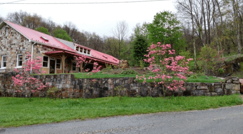 A stone house with a red roof surrounded by blooming pink flowers and green trees on a grassy landscape.