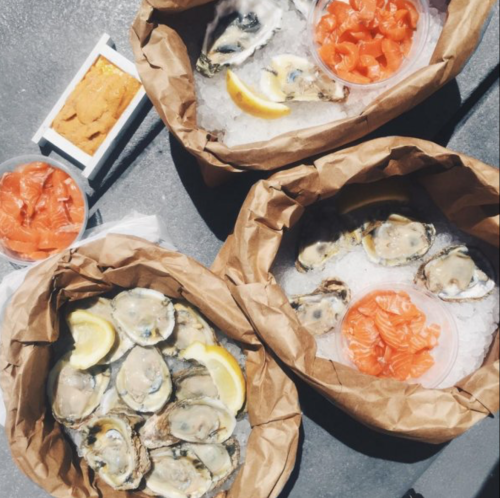 Three containers of fresh oysters on ice, garnished with lemon slices, alongside bowls of salmon and sea urchin.