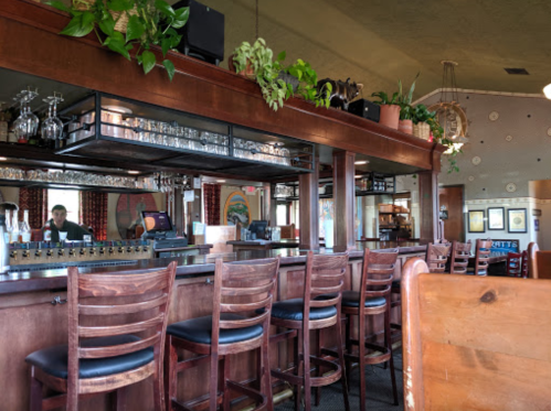 A cozy restaurant bar with wooden stools, plants, and a mirror reflecting the bartender in the background.