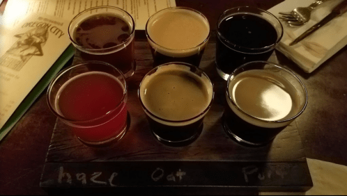 A wooden flight board with seven small glasses of different craft beers, labeled with their names.
