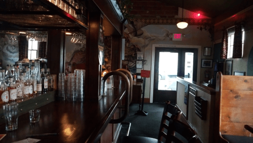 A cozy bar interior with wooden furnishings, shelves of liquor, and a door leading outside. Warm lighting and decor visible.