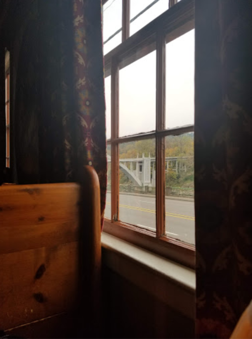 View from a window with wooden frame, showing a road and a bridge in the distance, framed by patterned curtains.