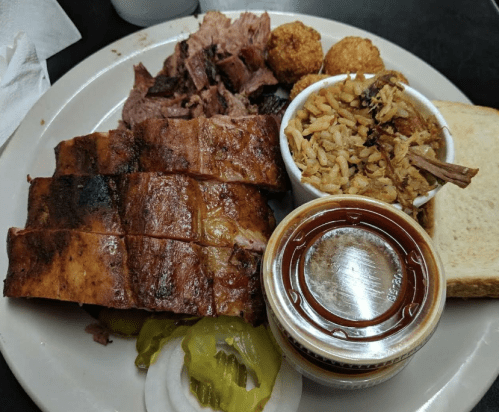 A plate of barbecue featuring sliced meat, rice, cornbread, pickles, onions, and a side of sauce.