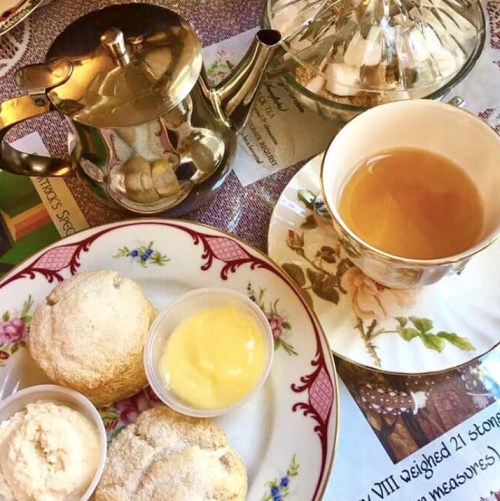 A silver teapot, a cup of tea, and two scones with cream and lemon curd on a floral plate.