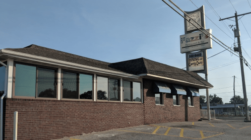 Exterior view of a brick restaurant with a sign, featuring large windows and a parking area. Clear blue sky above.