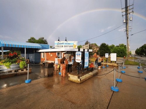 A colorful frozen custard stand with a rainbow overhead, people enjoying treats outside on a rainy day.
