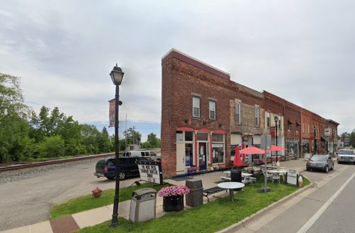 A small town street featuring brick buildings, outdoor seating, and a nearby railroad track under a cloudy sky.