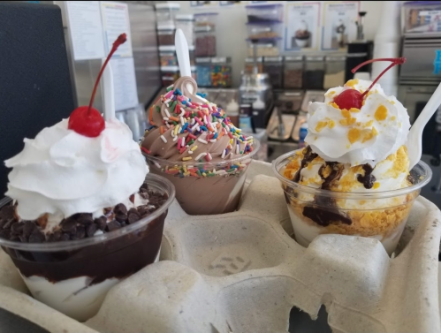 Three ice cream desserts in cups: one with chocolate, one with sprinkles, and one with crushed cookies, all topped with whipped cream and cherries.