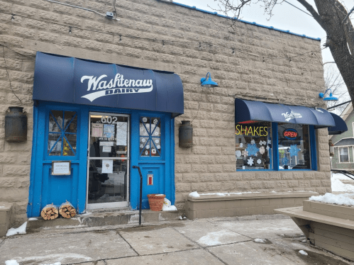 Exterior of Washtenaw Dairy with blue awnings, open sign, and snow on the ground. Cozy, inviting atmosphere.