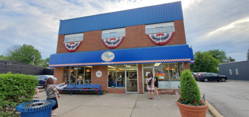 A brick storefront with blue awning and patriotic decorations, featuring people outside and a bench in front.