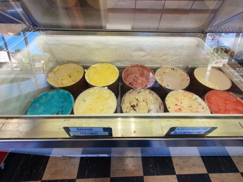 A display of various ice cream flavors in tubs, including banana, coconut, and blue raspberry, in a shop setting.
