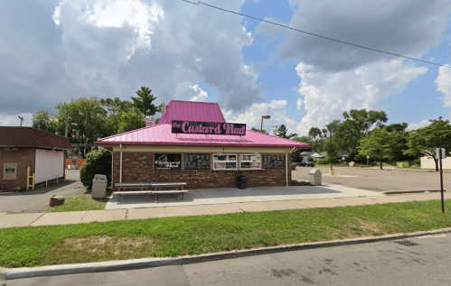 A small brick building with a pink roof, featuring a sign that reads "the Custard Hut" and outdoor seating.