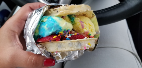 A hand holding a colorful ice cream sandwich with sprinkles and strawberries, inside a car.