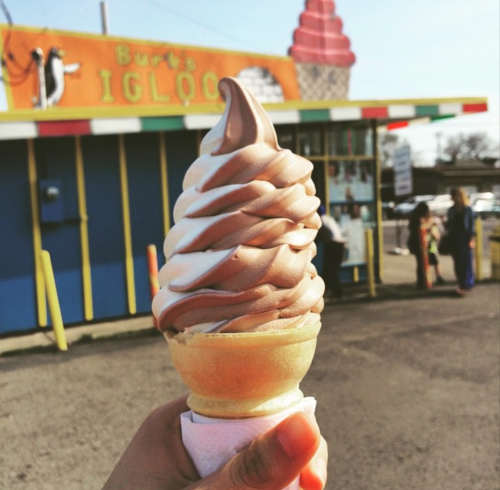 A hand holds a swirled chocolate and vanilla soft serve ice cream cone in front of Burt's Igloo ice cream shop.