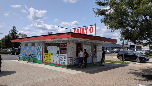 A colorful ice cream stand named "Dairy-O" with people ordering, surrounded by parked cars and bicycles.
