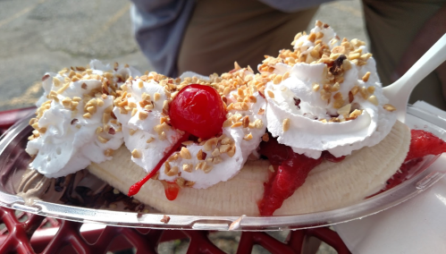 A banana split topped with whipped cream, cherries, and chopped nuts, served in a clear plastic dish.