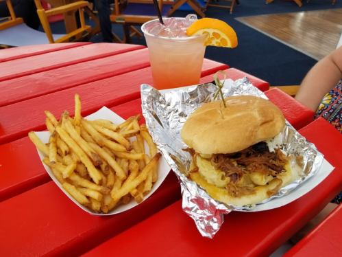 A plate with a pulled pork sandwich, crispy fries, and a colorful drink with an orange slice on a red table.