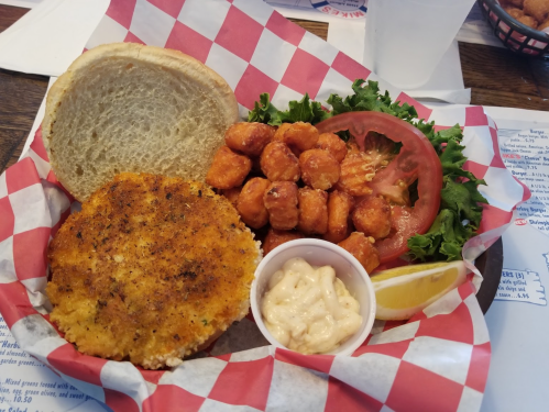A plate featuring a breaded chicken patty, tater tots, lettuce, tomato, a lemon wedge, and a side of creamy sauce.
