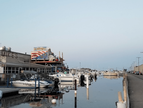 A calm marina with boats docked, reflecting buildings and a mural in the water under a clear sky.