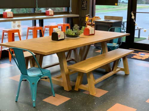 A wooden table with a bench and colorful chairs in a casual dining space, with large windows and a view outside.