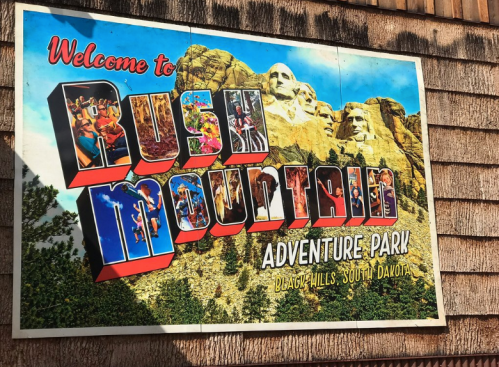 Colorful sign for Rush Mountain Adventure Park, featuring images of attractions and Mount Rushmore in the background.