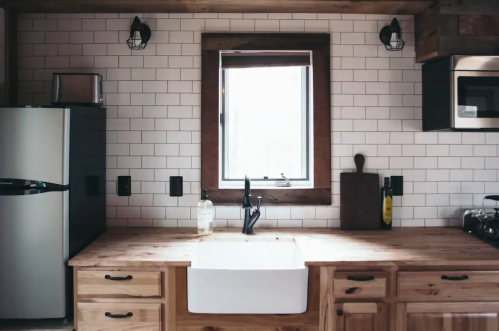A modern kitchen with a farmhouse sink, wooden cabinets, and a window framed in wood, featuring tile backsplash and appliances.