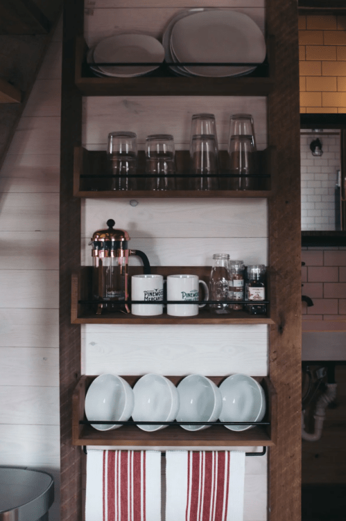 A wooden shelf displaying glasses, mugs, a French press, and white bowls in a cozy kitchen setting.