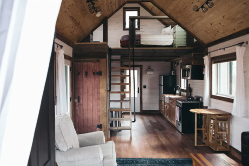 Cozy interior of a tiny house featuring a lofted bed, kitchen, and wooden staircase. Warm, rustic decor throughout.
