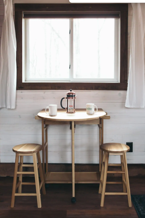 A small wooden table with two stools, a French press, and two mugs, set by a window with white curtains.