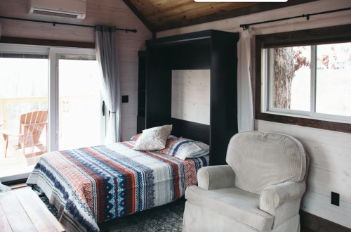 Cozy interior of a cabin featuring a bed with a patterned blanket and a comfortable armchair by a large window.