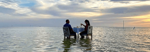 A couple dining at a table in shallow water during sunset, with a serene sky and calm ocean in the background.