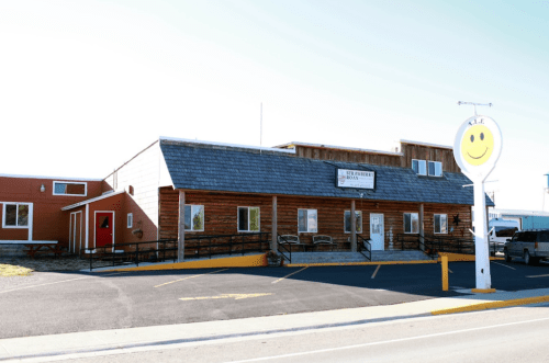 A wooden building with a sign and a smiling face, featuring a parking area and a clear blue sky.
