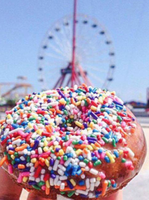 A hand holds a colorful sprinkle donut with a ferris wheel in the background on a sunny day.