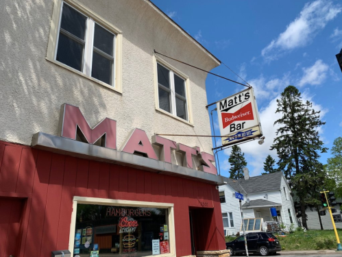 Sign for Matt's Bar with "Budweiser" logo, featuring a red and white color scheme, set against a blue sky.