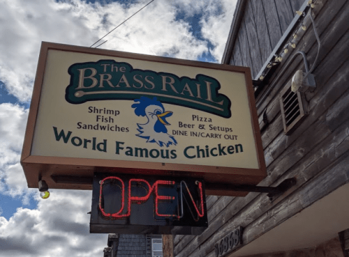 Sign for "The Brass Rail" featuring shrimp, fish, sandwiches, pizza, and "World Famous Chicken," with an "Open" neon sign.