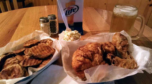 A table with fried chicken, coleslaw, and potato wedges, alongside a mug of beer and a beer menu.
