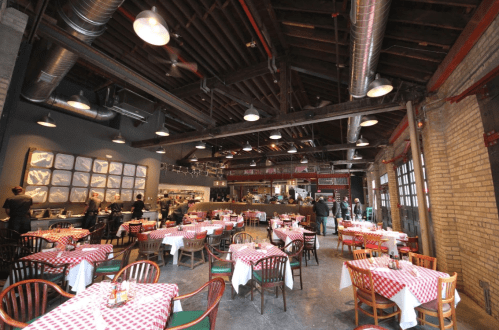 A spacious restaurant interior with checkered tablecloths, wooden chairs, and a bustling kitchen in the background.