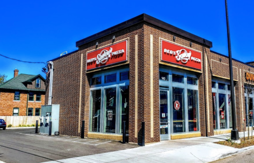 Exterior of Reed's Pizza, a brick building with large windows and a red sign, on a sunny day.