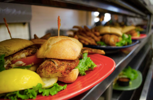 A close-up of a sandwich with fried chicken, lettuce, tomato, and lemon, served on a red plate with fries in the background.